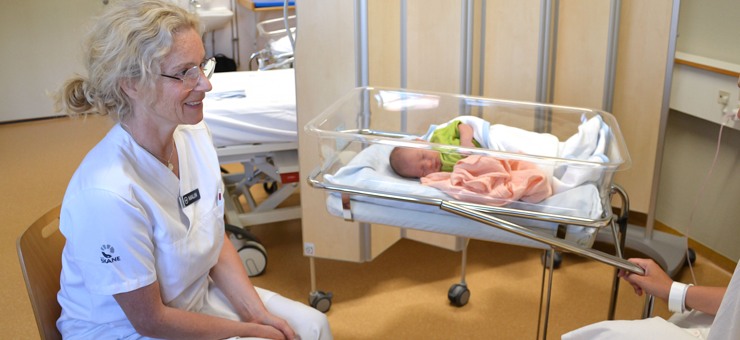 En barnmorska sitter jämte en bebis som sover i en säng och pratar med den nyblivna mamman.