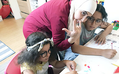 Föräldrastödjare Ghufran står lutad över ett skrivbord med var sin dotter sittandes bredvid. Ghufran hjälper sina döttrar med läxorna. Hon tecknar "I love you" på amerikanskt teckenspråk.