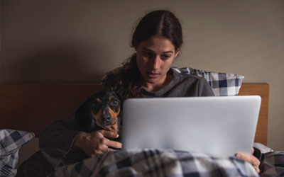 Person som är nerbäddad i en säng tillsammans med en hund. Personen har en dator i knäet och ser förkyld ut.