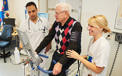 en patient cyklar på en motionscykel medan två sjuksköterskor står bredvid.