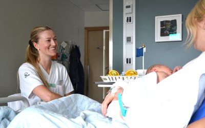 En sjuksköterska ler mot en nybliven mamma som håller sin bebis i famnen. 