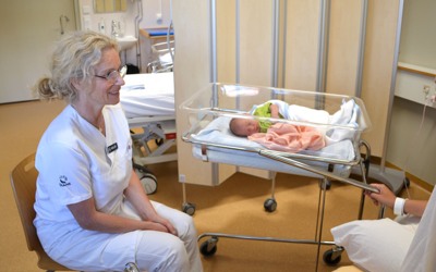 En barnmorska sitter jämte en bebis som sover i en säng och pratar med den nyblivna mamman.